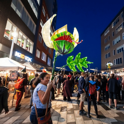 Une femme souriante brandit une lanterne géante illuminée en forme de plante carnivore, au milieu d’une foule rassemblée dans une rue piétonne en soirée, lors de Rivières de Lumières 2024.