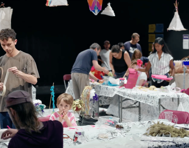 Atelier de fabrication de lanternes en papier et en osier : des adultes et un enfant assemblent des structures à des tables, sous des lanternes suspendues en forme de tipis et de cerfs-volants.