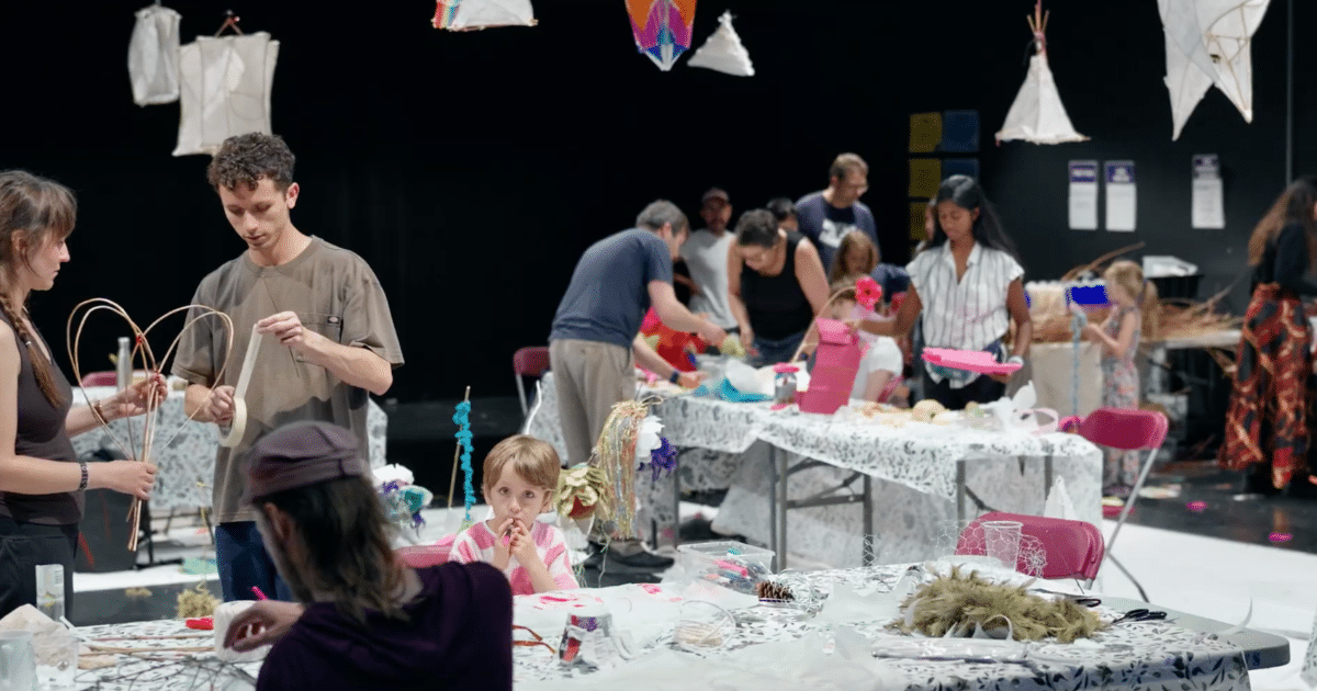 Atelier de fabrication de lanternes en papier et en osier : des adultes et un enfant assemblent des structures à des tables, sous des lanternes suspendues en forme de tipis et de cerfs-volants.