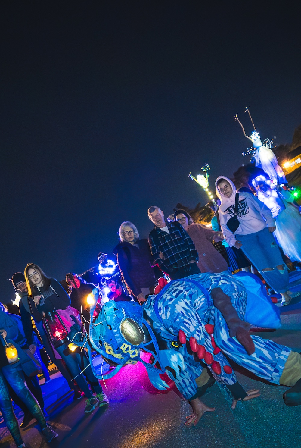 Une créature géante aux teintes bleues et lumineuses domine la scène, sous les regards d'une foule tenant lanternes et fanaux, lors du déambulatoire nocturne de Rivières de Lumières.