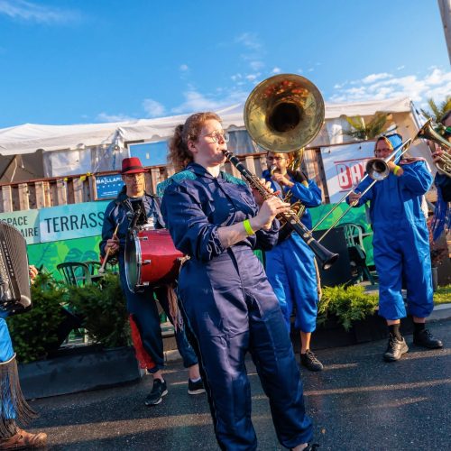 Photo de la fanfare Bungalow en pleine performance extérieure en plein jour : sept musiciens en combinaisons bleues à franges jouent en marchant dans une rue, avec accordéon, tambour, clarinette, trombones, sousaphone et trompette, devant une terrasse et un chapiteau blanc.