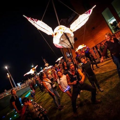 Photo nocturne de la parade des lanternes : Xavier Graveline porte sur une perche une grande lanterne lumineuse en forme de tête d'animal ailé (oreilles ou ailes pointues), tandis qu'une file de participants et de spectateurs avance le long d'un bâtiment de brique éclairé, sous un ciel noir.